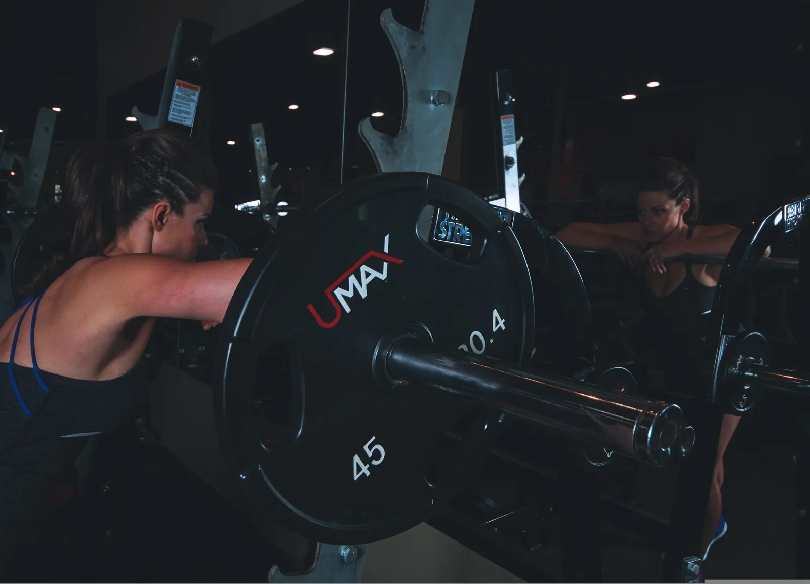 A woman resting her arms on the barbell in the rack, in a dark gym